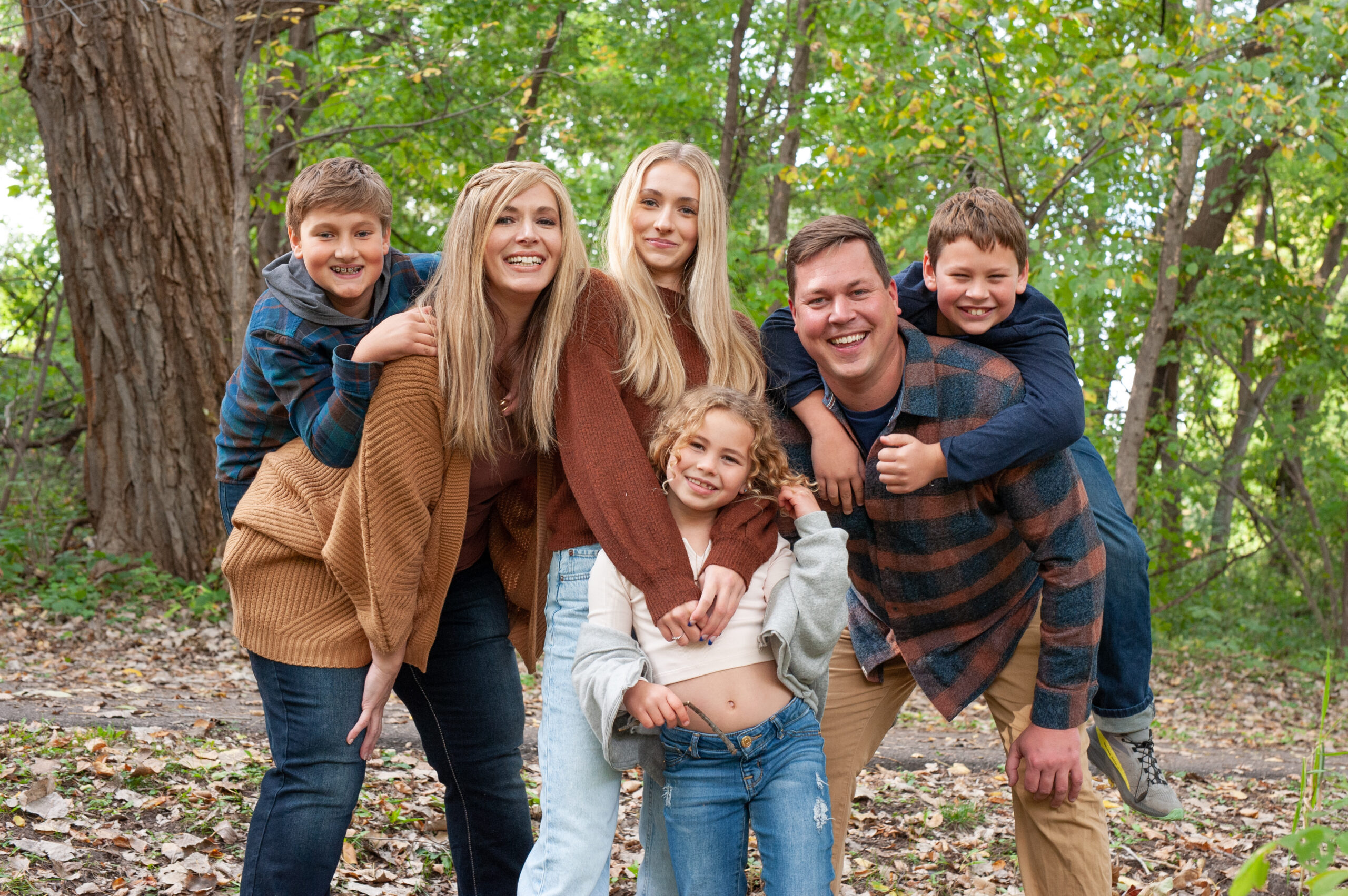 Set on a path in the woods as the green leaves are just starting to turn to golds and reds, a blended family of six are all leaning in close and happy for a casual, cozy portrait. The group includes two boys, two girls, a mom and dad. The two boys are getting piggy back rides from the parents on each end.